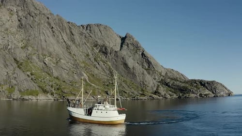 Aerial drone shot of fishing boat on the ocean with huge rock faces and mountains in the background
