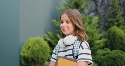 Side view of smiling pretty schoolgirl with school bag and books walking in the street