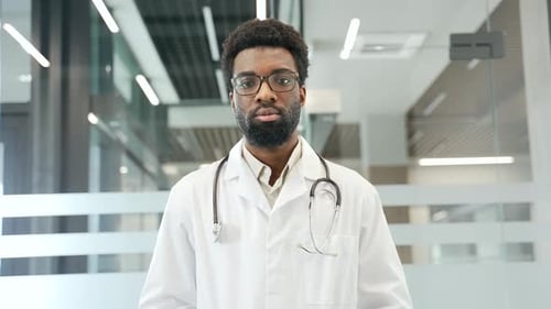 Confident african american male doctor standing in modern medical office, wearing white coat