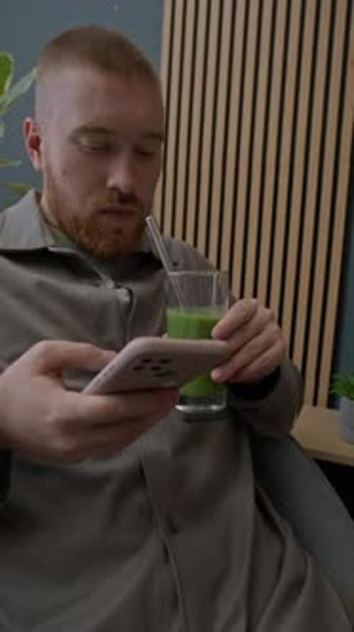 Young Man Drinking Green Smoothie while Scrolling Through Phone