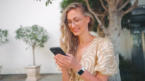 Smiling Young Woman Using Smartphone Near Plants in City