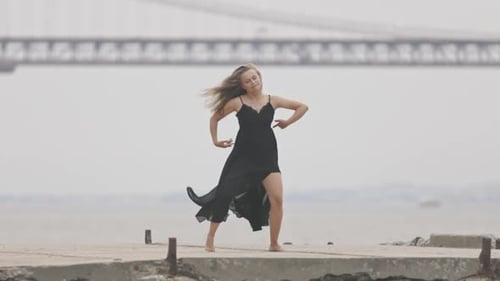 An Adult Woman in Black Dress Gracefully Dancing on the Pier in Slow Motion with a Bridge on the