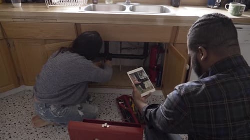 Woman Repairing Sink While Man Reads Tablet