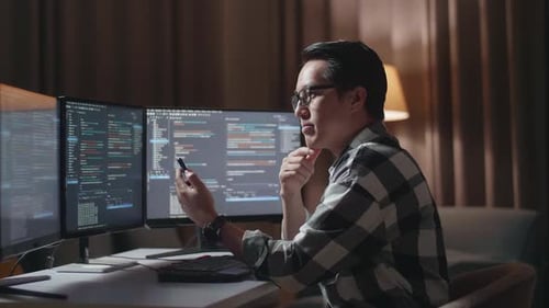 Asiatischer Programmierer schaut auf den Desktop-Computerbildschirm und benutzt das Smartphone, während er Software erstellt