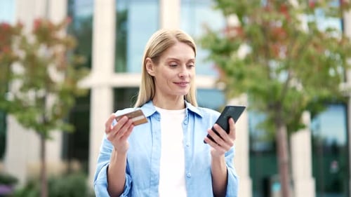 Smiling female is shopping online using credit card and phone standing on the city street near