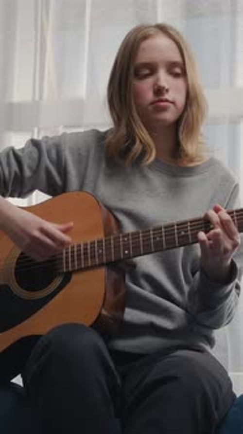 Thoughtful Young Musician Practicing Acoustic Guitar at Home Near Bright Window Focusing on Gentle