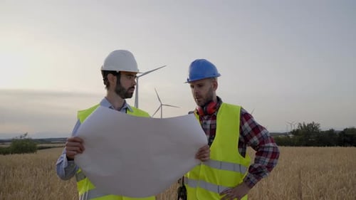 Two Engineers in Hard Hats Work Looking at a Blueprint in an Agricultural Field with Wind Turbines