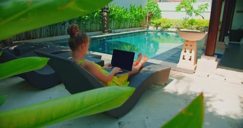 Woman uses laptop, lying on a lounger near to swimming pool. Freelancer, remote work. Summer vacatio