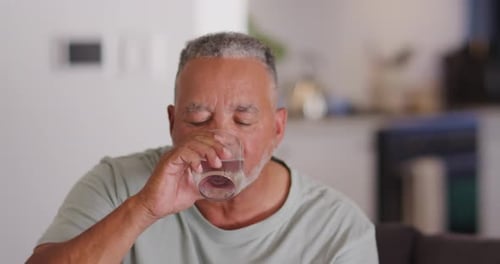Älterer afrikanischer Mann hat Durst, hebt klares Glas, trinkt langsam auf dem Sofa zu Hause und lächelt