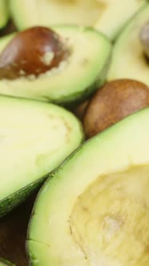 Vertical video. Numerous avocado halves with pits have taken over the entire space on the table.