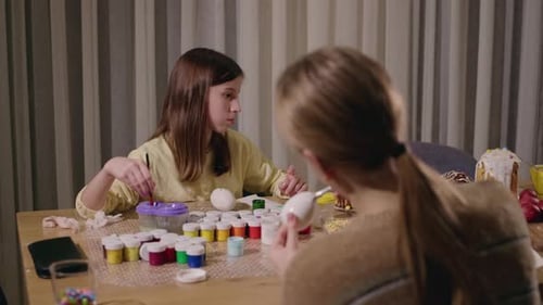 Two Teenage Girls Painting Easter Eggs at a Table Filled with Colorful Paints Brushes and Festive