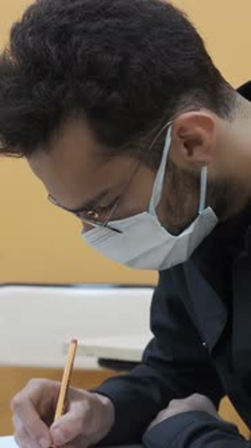 Masked Adult Writes on Paper in a Workplace