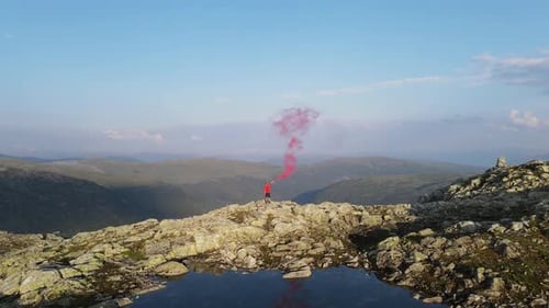 Hiker releasing red smoke on rocky peak with alpine lake and hills