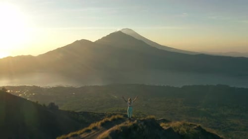 Female adventure hiker with raised arms standing on ridge looking at scenic Bali mountain landscape