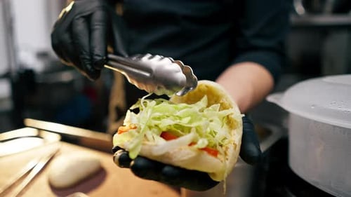 Close Up Chef in Black Gloves Puts Different Ingredients Into a Bun with Tongs