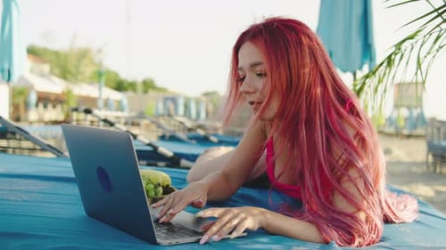 Junge Frau mit rosa Haaren arbeitet an einem sonnigen Tag in einem tropischen Resort am Laptop am Pool
