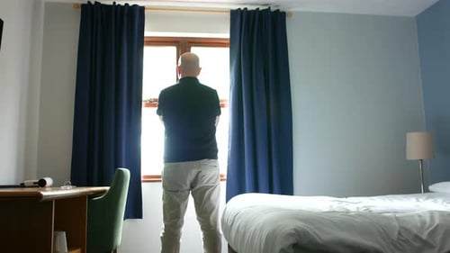 Man Stands in Hotel Room Looking Out Window