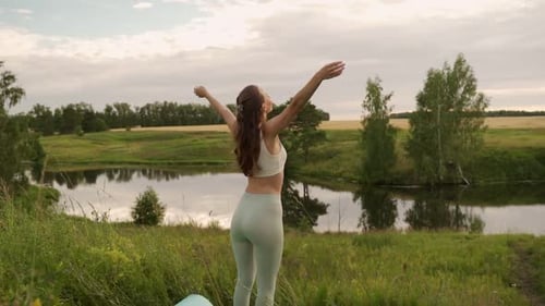 Young Woman Stretches Her Arms Up Doing Yoga Against the Background of a Pond