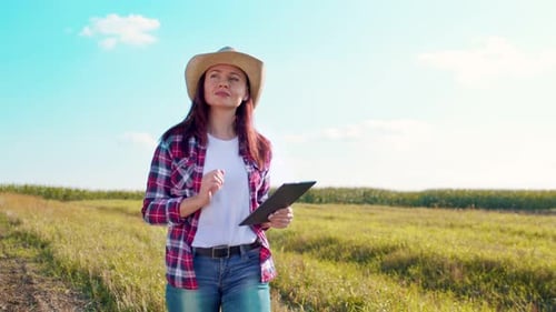 Caucasian Woman Farmer in a Hat Walking Through the Middle of the Golden Field of Wheat and Tapping