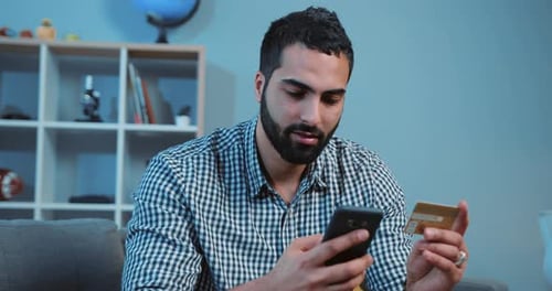 Close Up of Smiling Man Buying Online Uses a Phone and a Credit Card on Sofa Home Shopping Cellphone