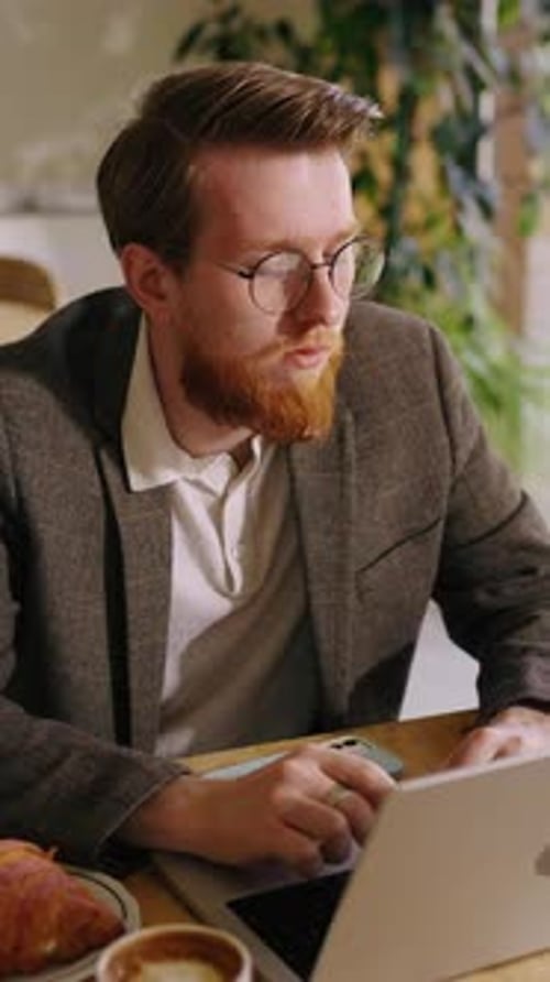Thoughtful Young Professional with Beard and Glasses Working on Laptop in Cozy Cafe Pausing to