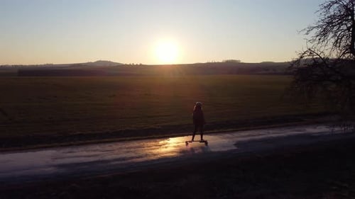 girl riding an electric longboard on a road surrounded by trees, at sunset in spring, aerial
