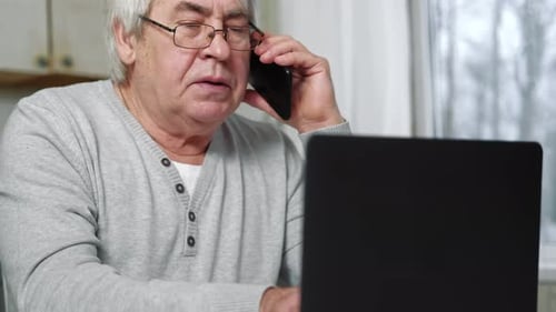 Seriöser älterer Mann mit Brille, sprechendes Handy, sitzt am Schreibtisch zu Hause am Arbeitsplatz mit