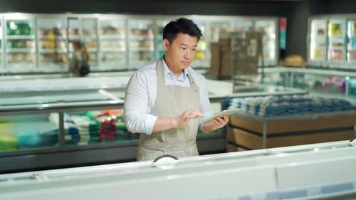 Asian grocery store manager salesman in apron using digital tablet counting goods in supermarket.