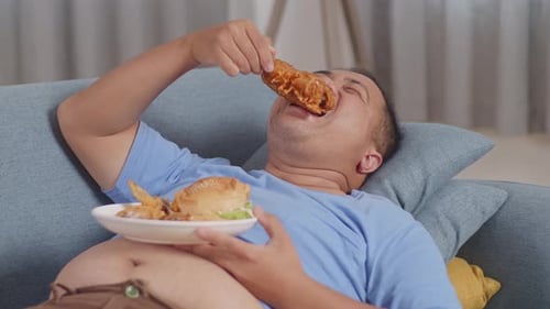 Close Up Of A Fat Asian Man Eating Fast Food Having Fried Chicken While Lying On A Sofa