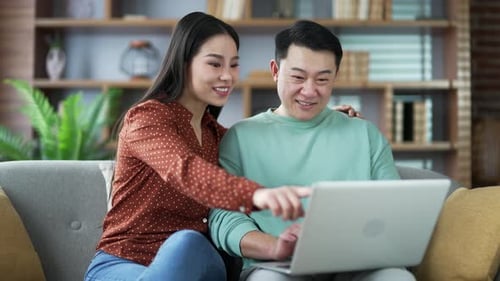 Happy asian couple shopping choosing products in online store using laptop, sitting on sofa