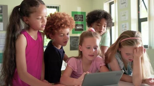 Schoolchildren leaning around tablet at classroom table, central girl touching screen to start game