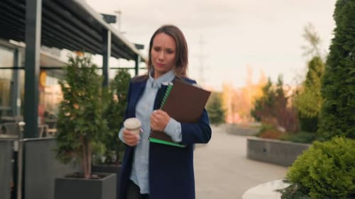 Woman Walks with Coffee and Notebook Outside Building