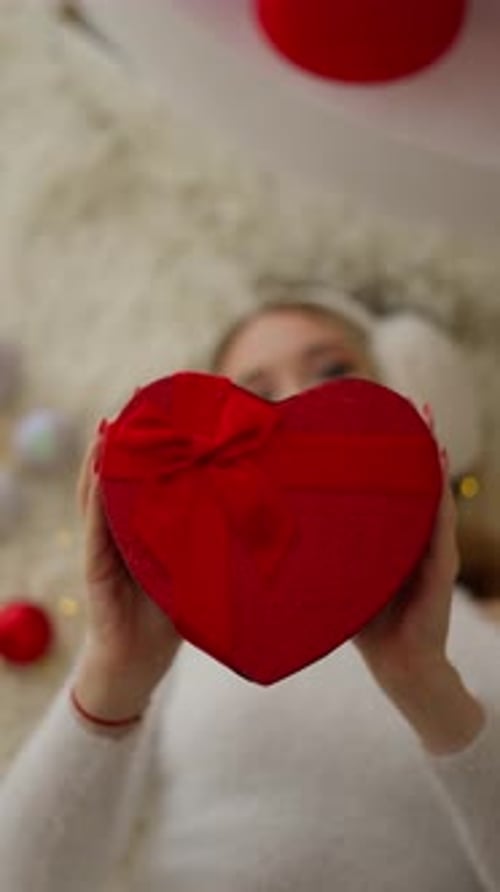 Woman Holds Heart Shaped Gift While Lying on Carpet