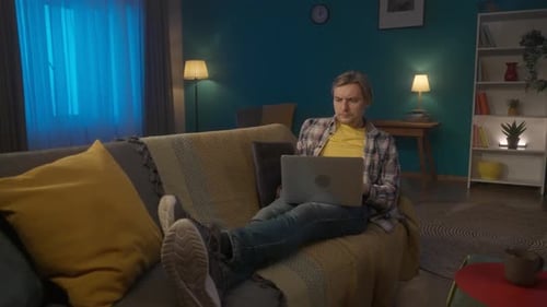 A Man is Lying on the Couch at Home with a Laptop on His Lap A Man with a Concentrated Face Works or