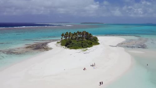 Low and slow aerial fly-over of small sandy island with palm trees and green vegetation. Camera pans