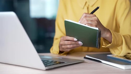 Close-up of businesswoman’s hands writing notes in notebook while using laptop at desk in modern