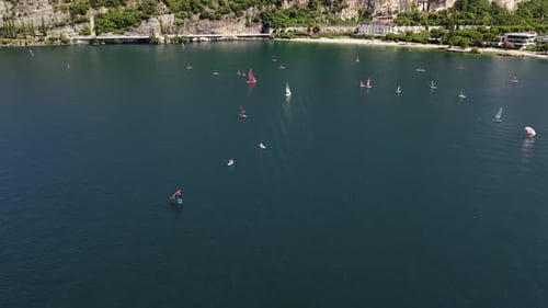Red catamaran and windsurfers on Lake Garda near Torbole with coastal hotel and cliffs, aerial flyov