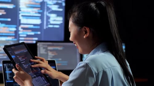 Back View Of Asian Female Programmer Looking At Database On Tablet While Writing Code By A Laptop