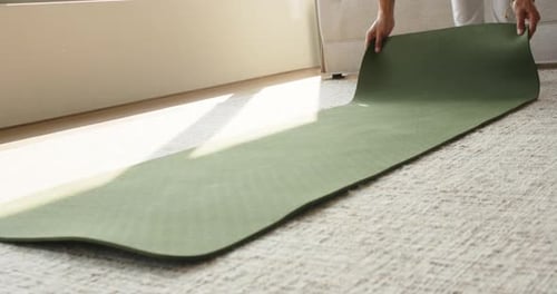 Person wearing sweats reaching and grabbing rolled green mat unrolling on beige carpet for workout
