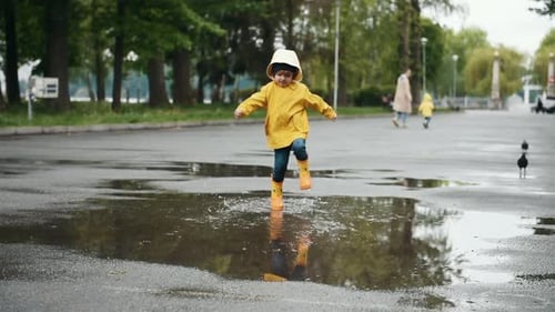 Kid in yellow waterproof cloak and boots running and jumping into puddle outdoors after the rain