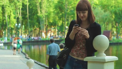 The girl reads the message on the phone while standing near the lake in the park