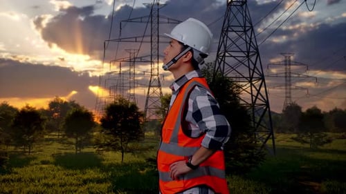 Side View Of Asian Male Engineer Looking Around With Arms Akimbo Near High Voltage Tower