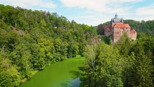 Historic castle perched on a hill surrounded by dense green forest and calm river, evoking a fairy-t