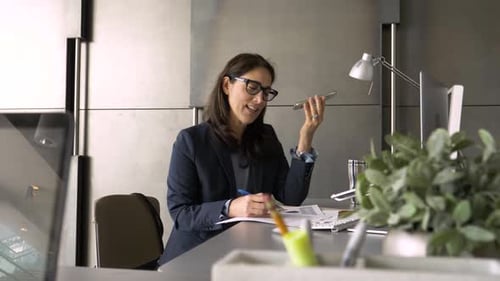 Mature businesswoman in suit talks on smart phone with paperwork at office desk