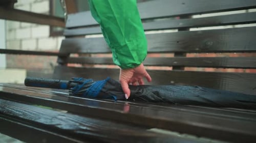 Individual in Green Raincoat Reaching for Umbrella on Swing Bench During Rainy Weather