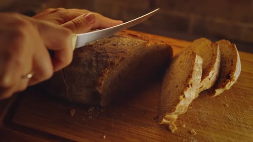 Close-up hands of female baker with knife cutting freshly baked aromatic loaf of wholemeal bread