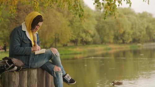 Young Creative Man with Dreadlocks Drawing Writing in Notebook Sitting on Stumps Near Lake in