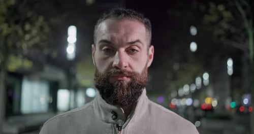 A Young Man with a Stylish Trendy Hairstyle and Beard in Casual Clothes Stands on a Night City