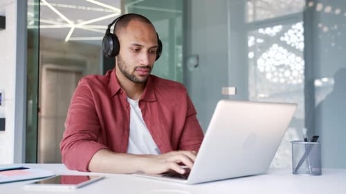 Focused businessman or IT specialist in wireless headphones works on a laptop sitting at a workplace