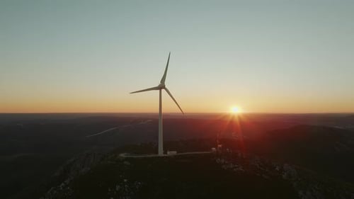 Aerial rotation of wind turbine on top of the mountain under blue and orange cloudless sky.